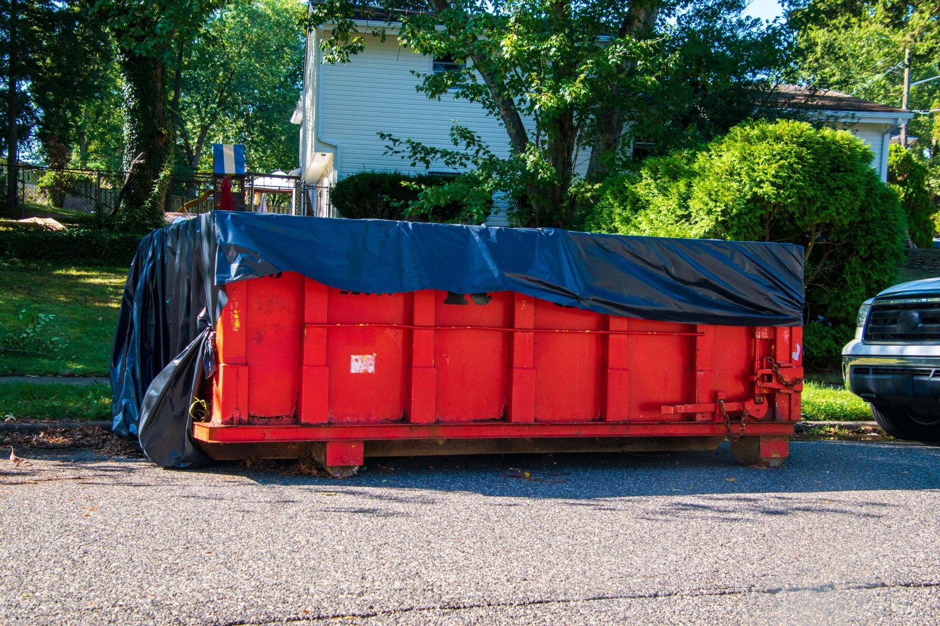 A large, bright red dumpster covered with a black tarp, parked on a gravel driveway in front of a house.