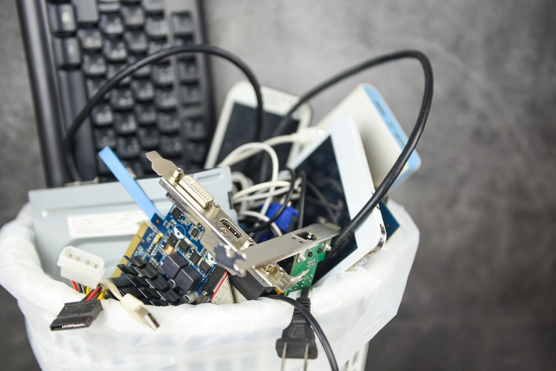 A white trash can filled with discarded electronics, including a keyboard, circuit boards, and tangled power cables.