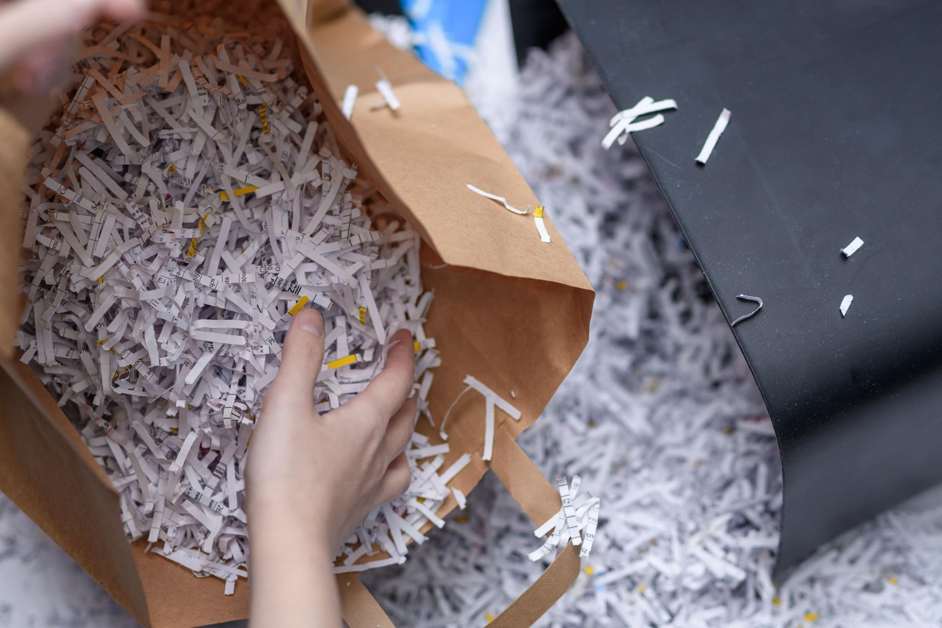 A hand reaches into a brown paper bag filled with shredded paper, with more shredded paper scattered on the floor.