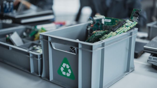 A grey plastic bin with a green recycling symbol contains electronic circuit boards, sitting on a table.