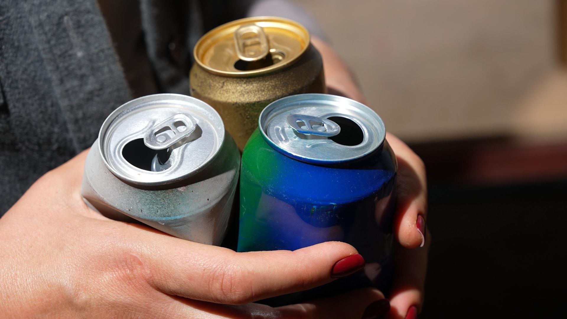 Hands with red-painted nails holding three open metal cans, one gold, one silver, and one blue and green.