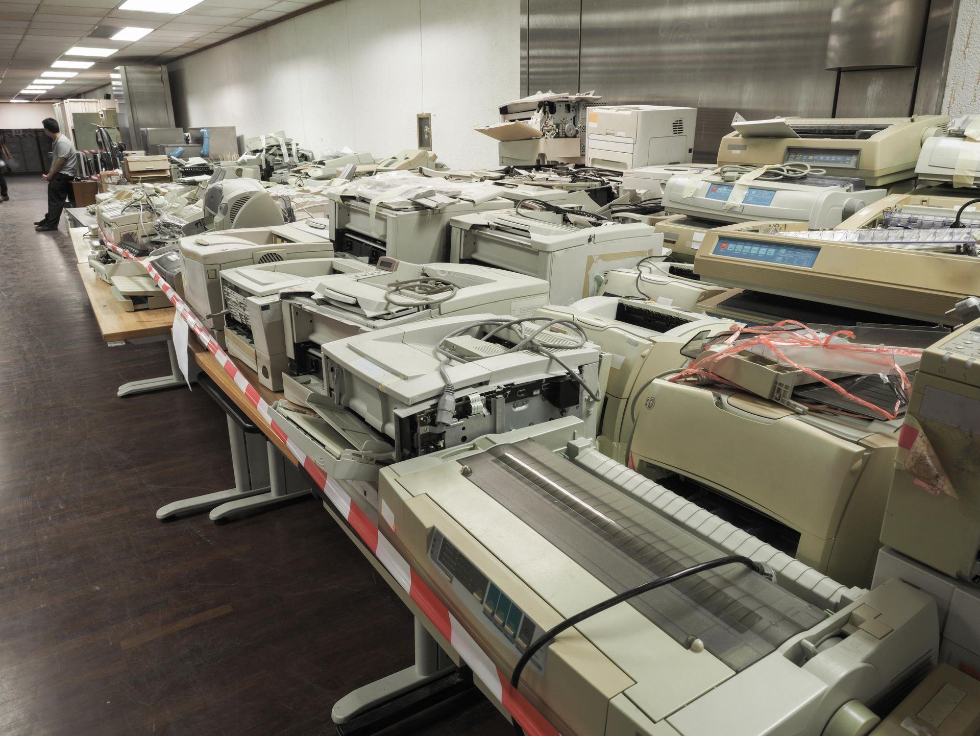 Rows of old printers and electronic equipment lined up on long tables in a warehouse or office space.