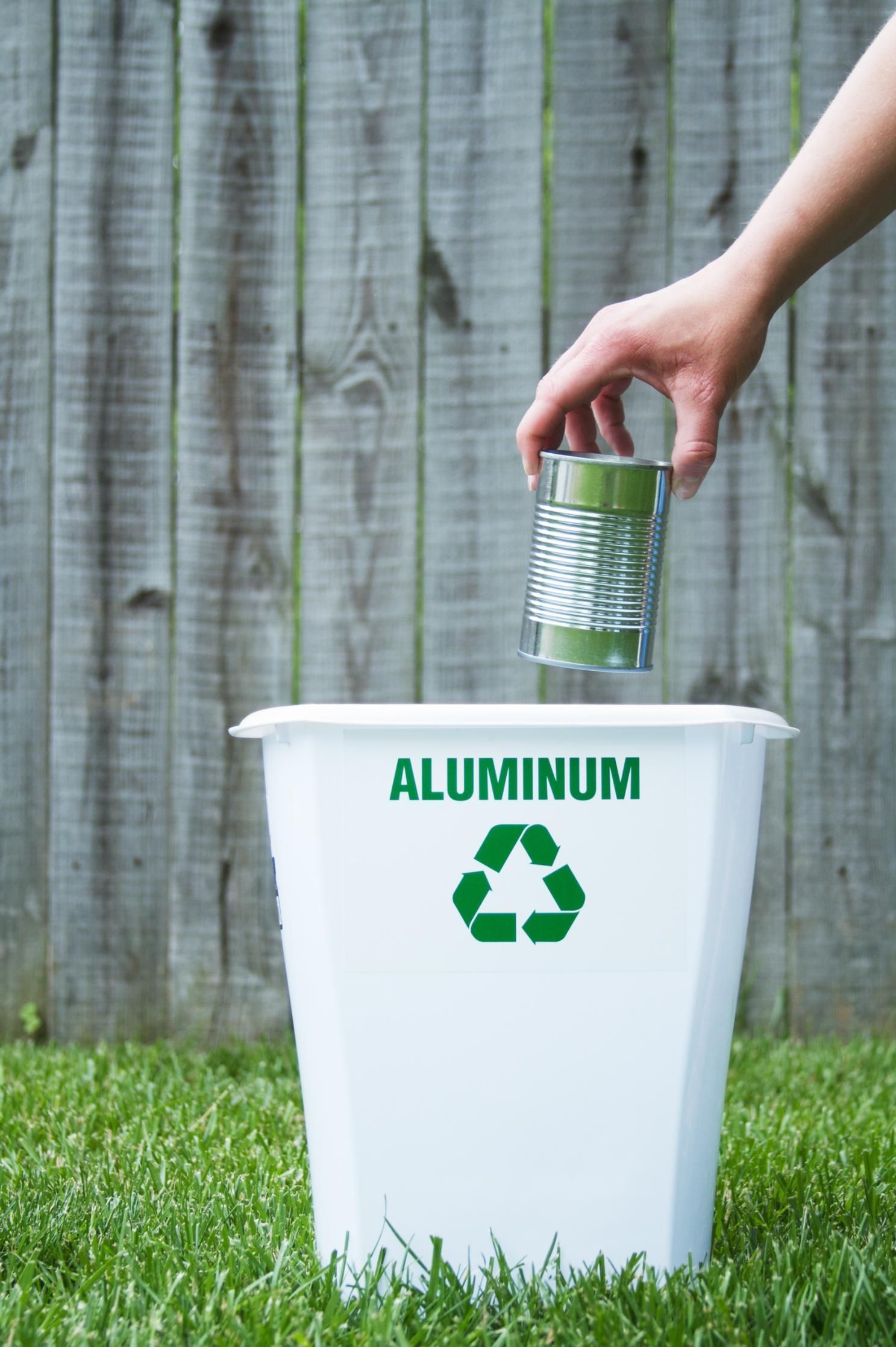 A hand placing an aluminum can into a white recycling bin labeled 