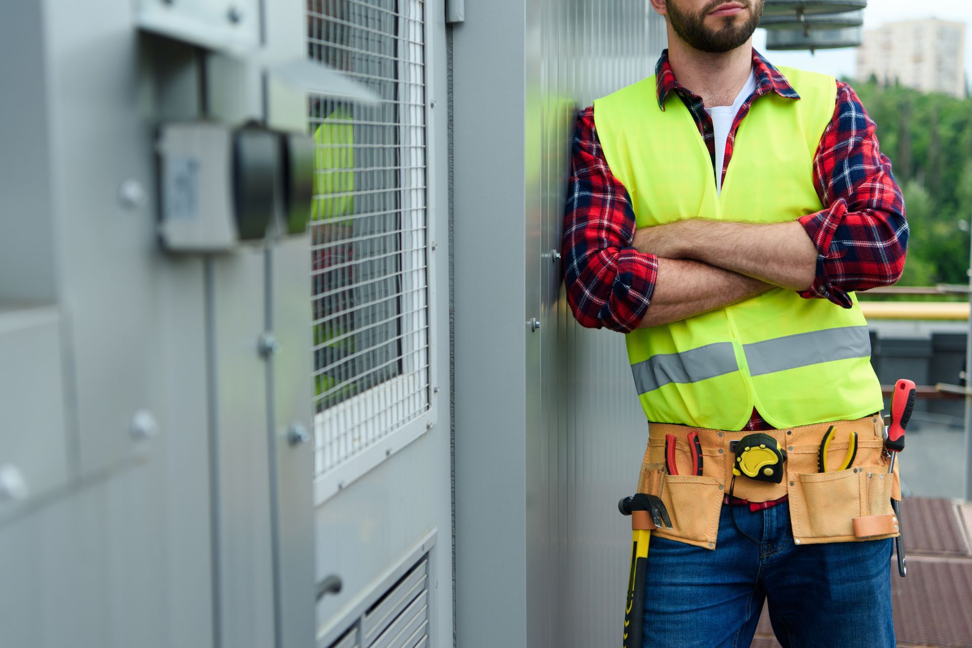 Construction worker leaning against a metal structure, arms crossed, wearing a yellow safety vest, and tool belt.
