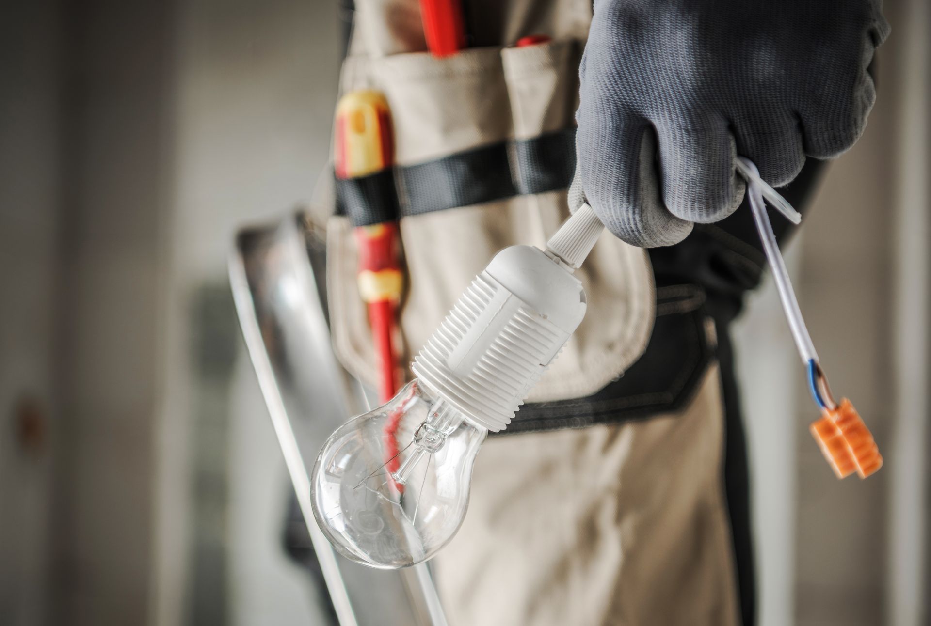 Electrician holding a lightbulb with wires, wearing gloves and tool belt.