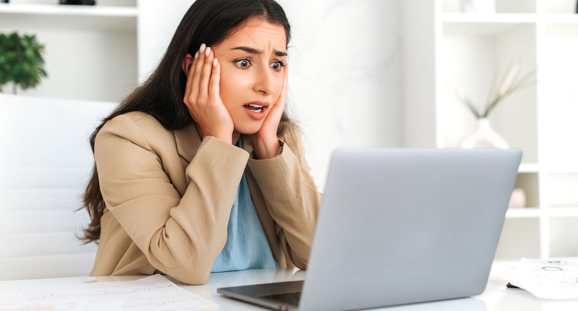 A woman is sitting at a desk looking shocked at a laptop computer.