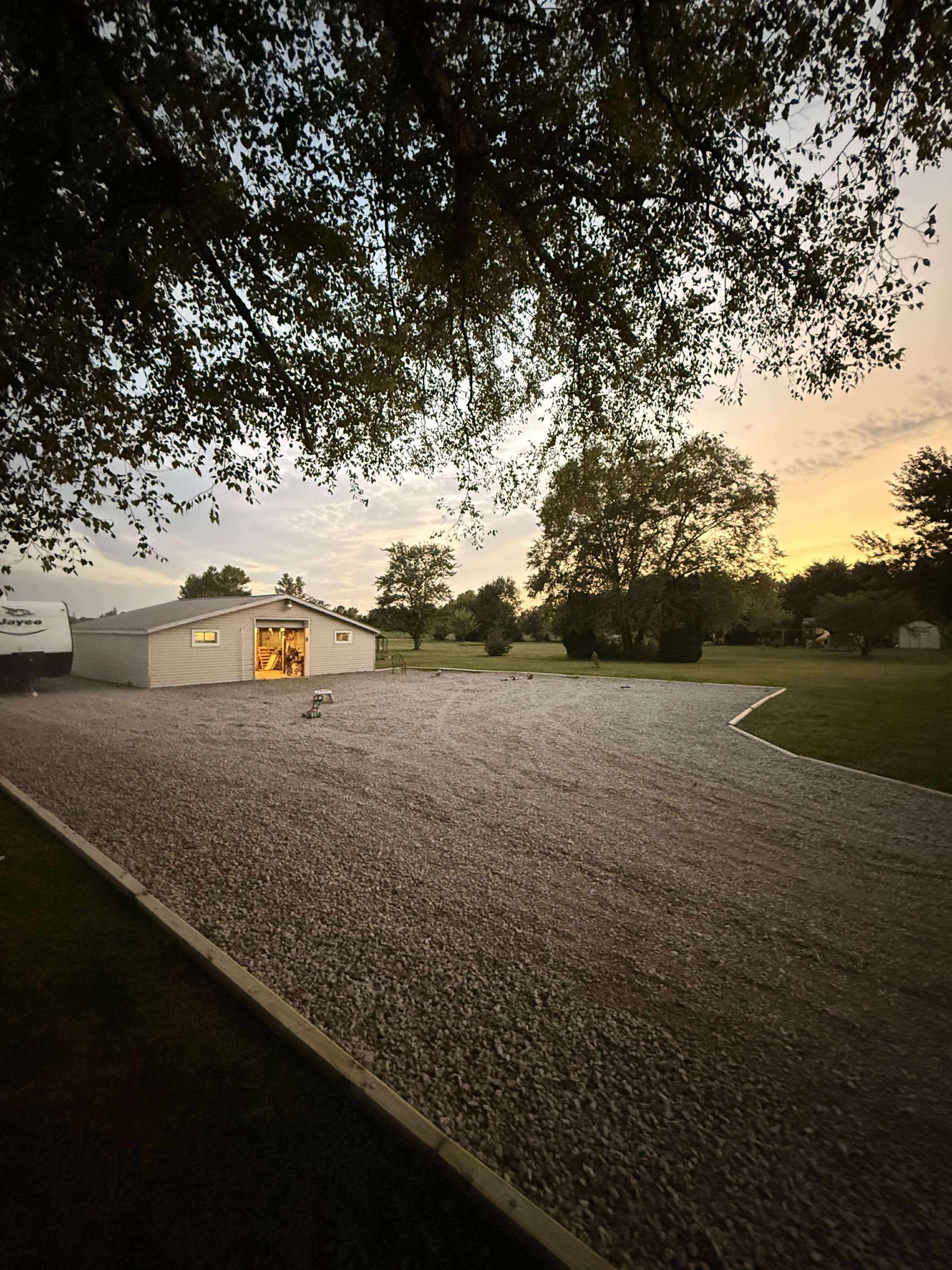 Gravel driveway leading to a white building at sunset. Silhouetted trees and colorful sky are in the background.