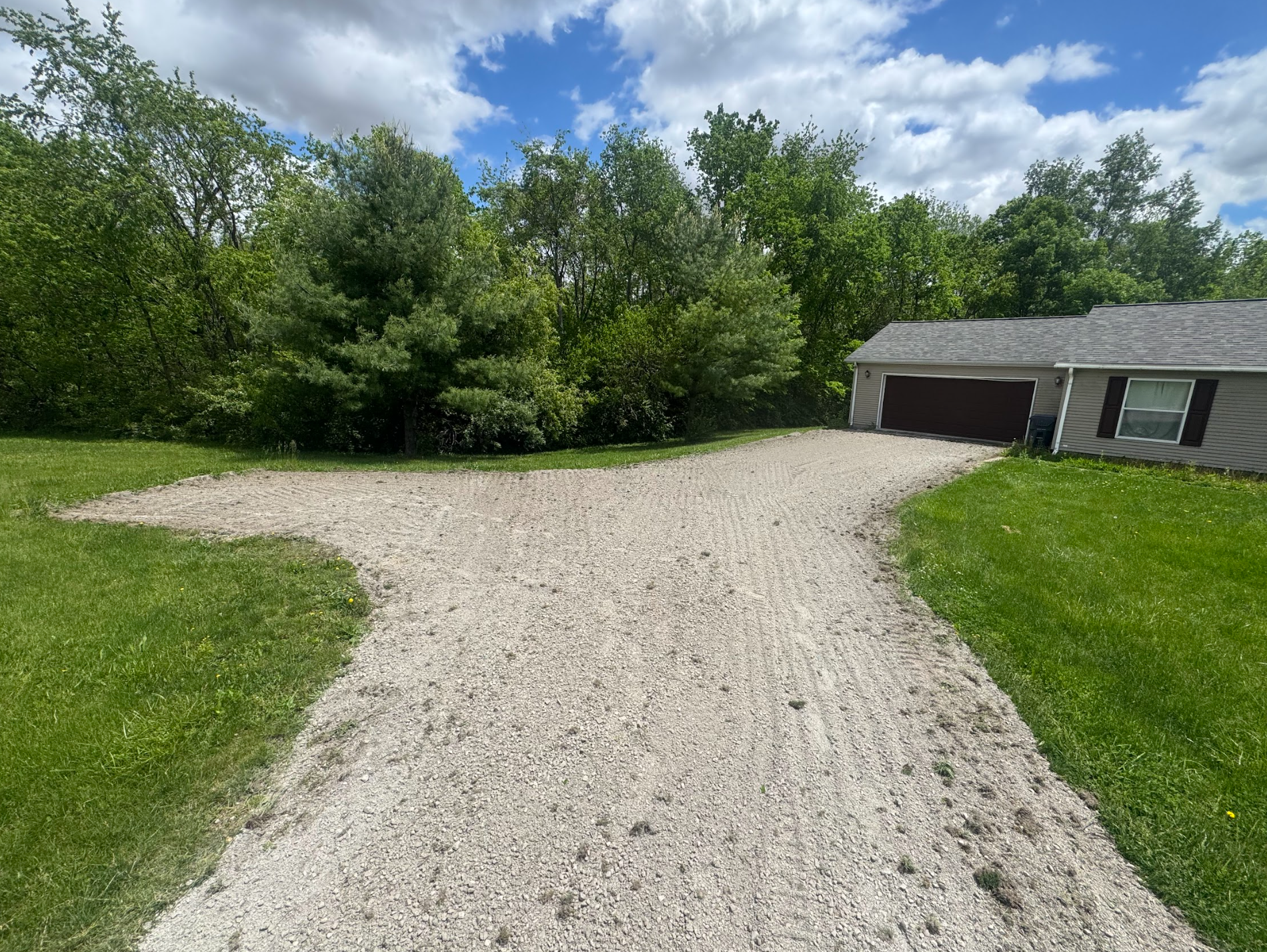 Gravel driveway leading to a garage and house, surrounded by green grass and trees under a blue sky.