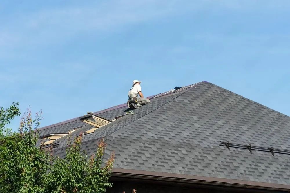 Roofer on a dark shingled roof, working under a blue sky.