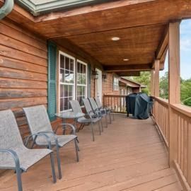 Covered wooden porch with chairs, a grill, and log cabin siding overlooking a scenic view.