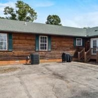 Single-story log cabin exterior with windows, porch steps, and trash bins beside a paved driveway.
