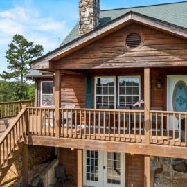 Wooden cabin with wraparound deck, stone chimney, and white-framed windows under a blue sky