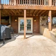 Covered concrete patio under a wooden deck with French doors, a grill on the left, and stairs on the right