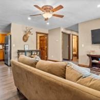 Cozy living room with beige sofa, ceiling fan, wall TV, and open doorway to kitchen and hall