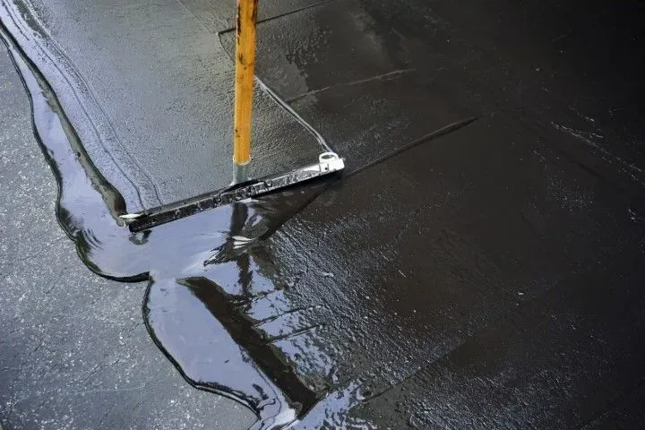 A squeegee with a yellow handle spreads a dark, viscous liquid sealant across a flat surface.