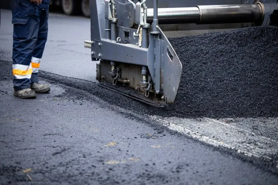 A road paver lays fresh black asphalt onto a road surface next to a worker wearing high-visibility work pants.