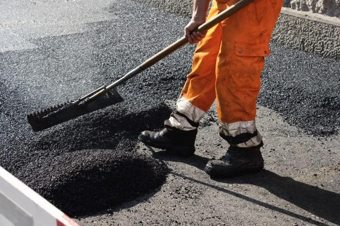 A road construction worker in orange overalls uses a tool to spread fresh, black asphalt on a road surface.