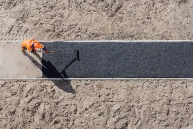 A worker in orange high-visibility gear uses a rake to spread fresh black asphalt on a narrow path.