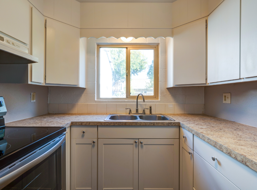 An empty kitchen with white cabinets , a stove , a sink and a window.