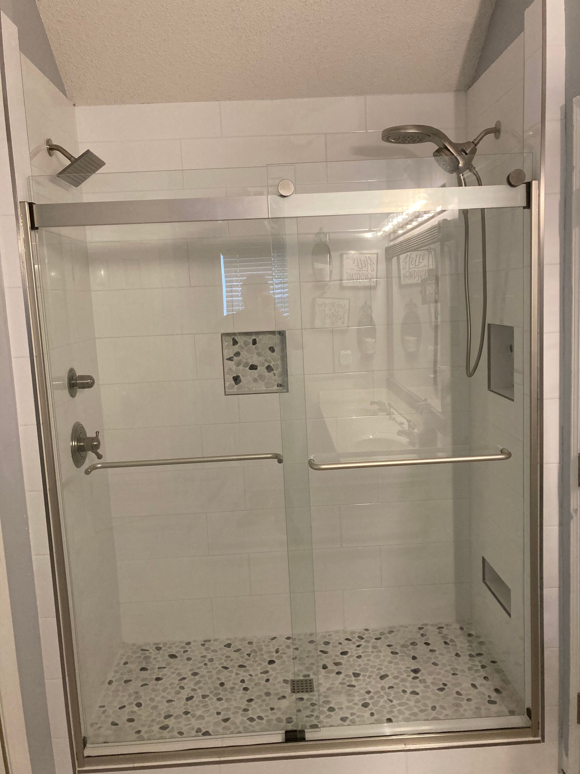 Modern glass shower enclosure with chrome hardware, white tiled walls, and a pebble-tiled floor.