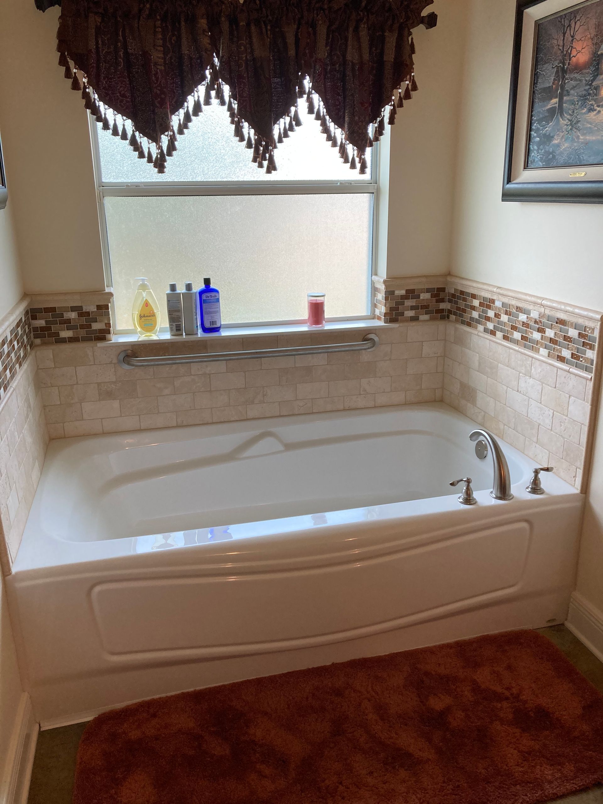 A white bathtub set against a wall with tan stone tile, a decorative border, a window, and a dark brown fabric valance.