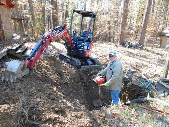 A man is standing next to a small excavator in the woods.