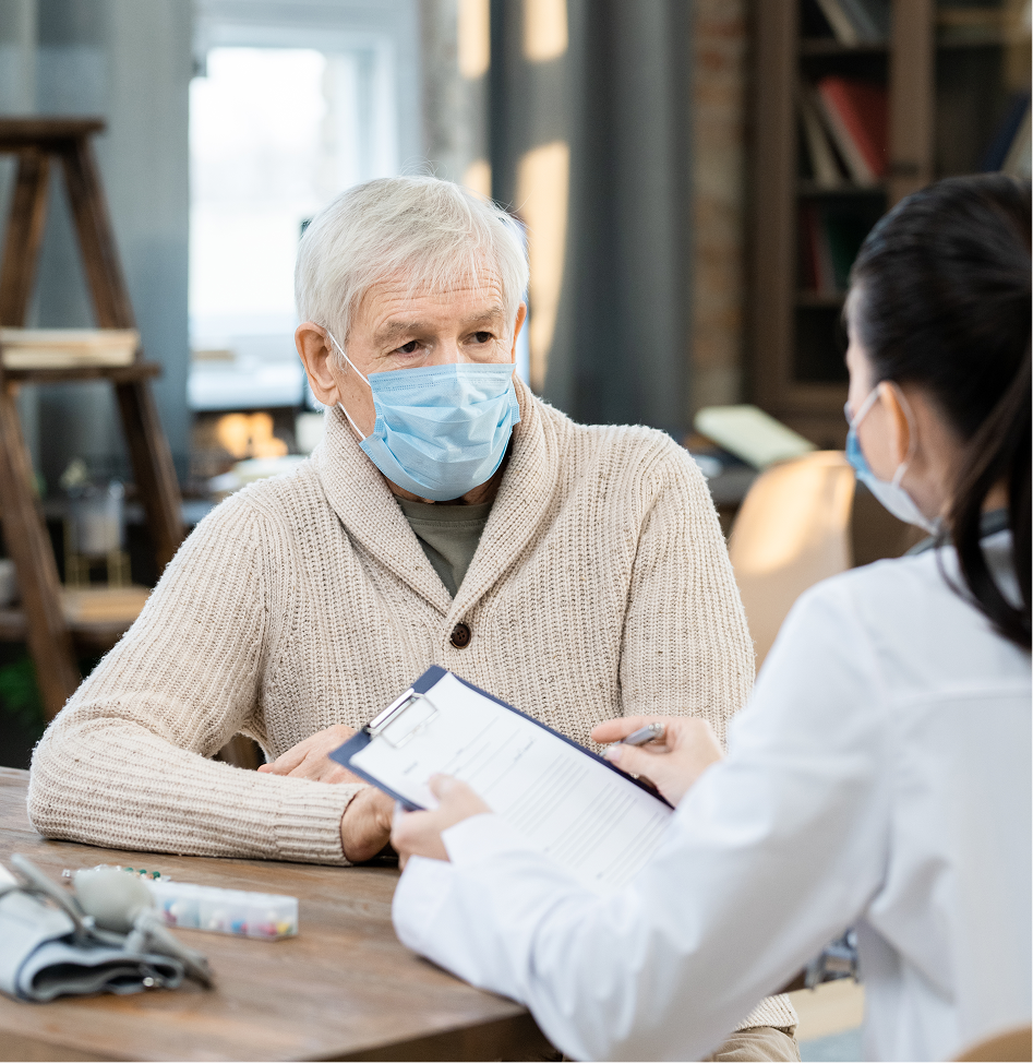 An elderly man wearing a mask is sitting at a table talking to a doctor.