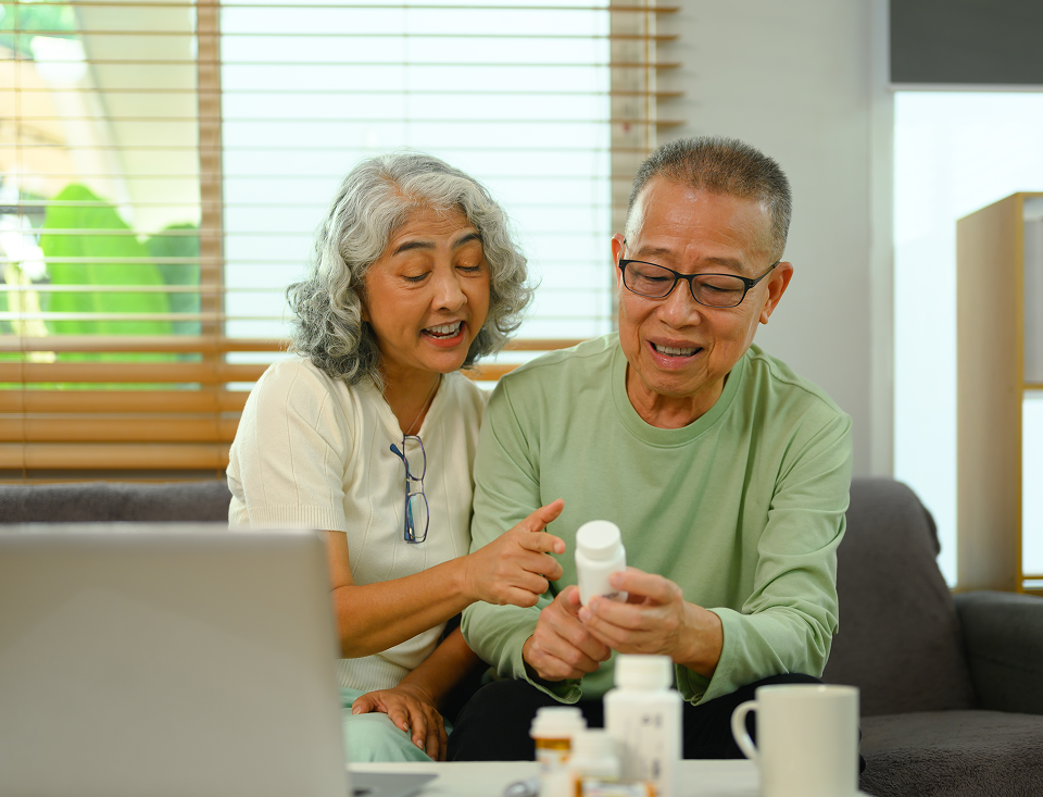 An elderly couple is sitting on a couch looking at a bottle of pills.