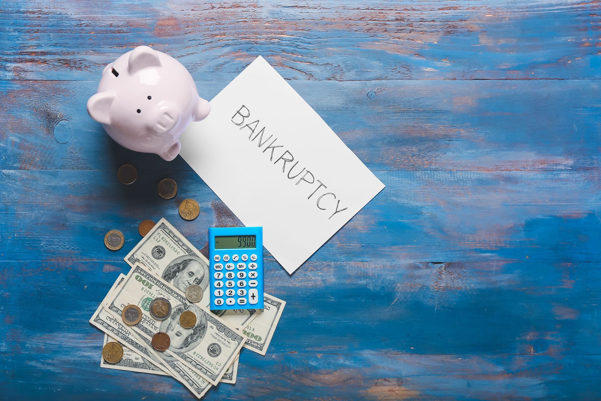 A piggy bank , calculator , money and a sign that says bankruptcy on a blue wooden table.