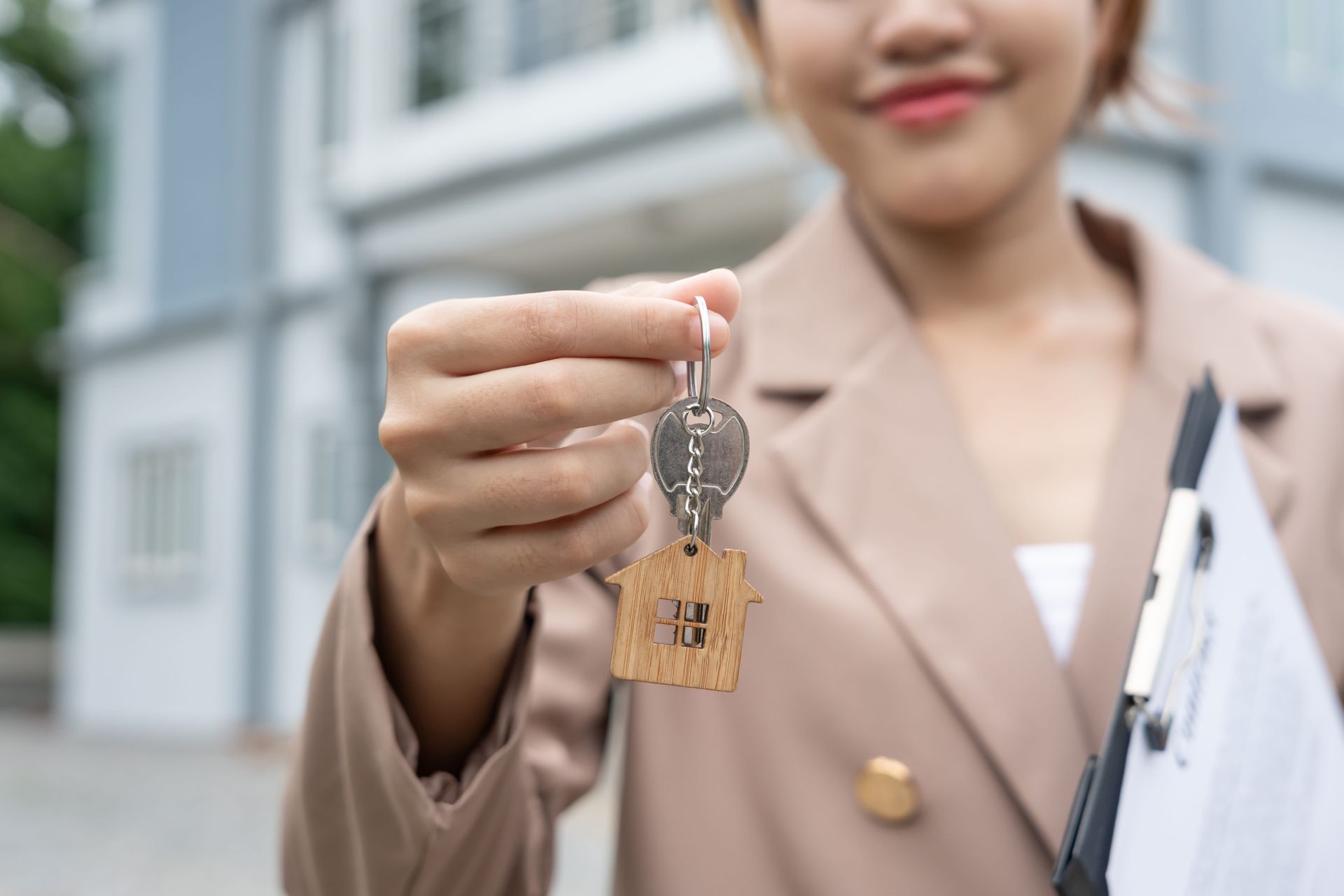 A smiling person in a tan blazer holds up a set of keys with a house-shaped wooden keychain in front of a building.