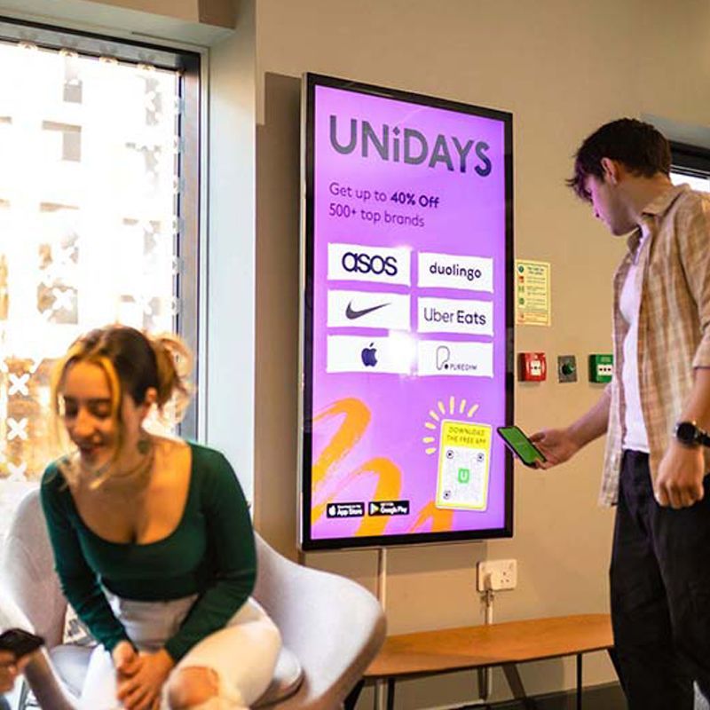 A man is holding a cell phone in front of a sign that says unidays