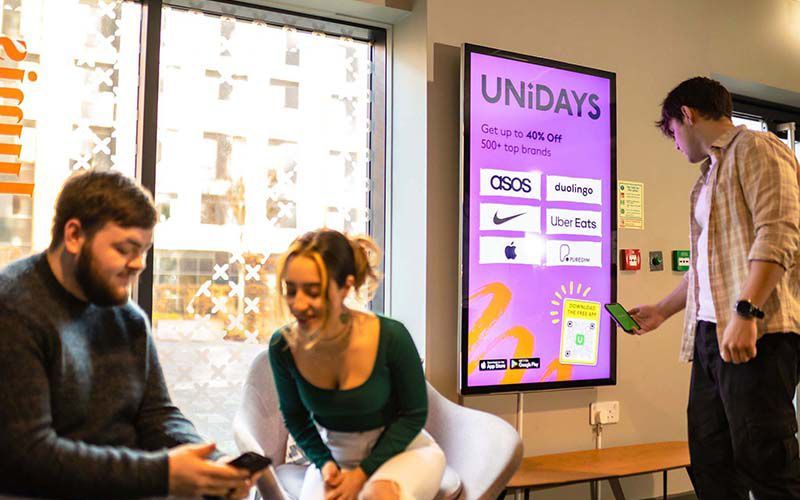 Three young adults interact near a UNiDAYS display. One scans a phone, two smile, sitting on chairs by a window.