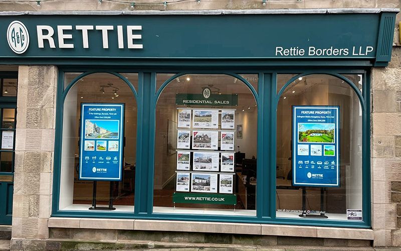 Exterior of Rettie estate agents shopfront with property listings. Teal and white signage on stone building.