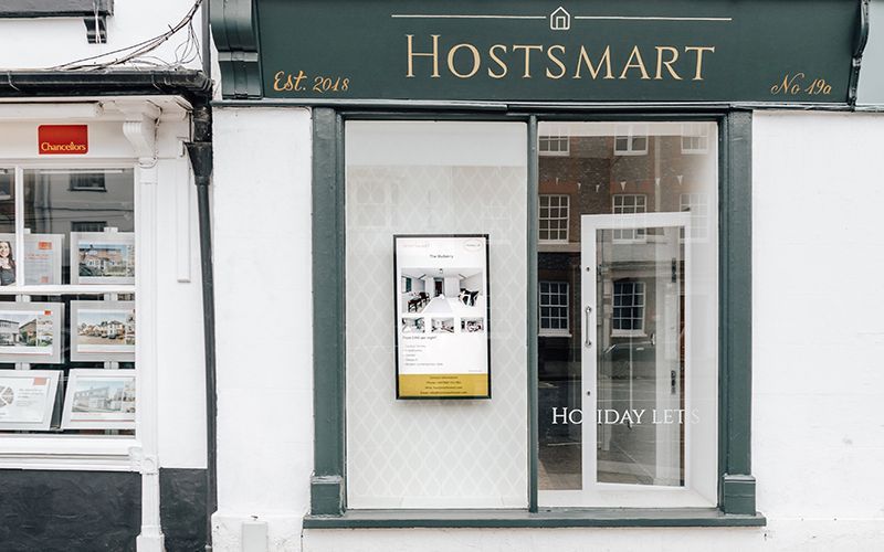 HostSmart storefront with digital display showing property photos; white building, green signage, window.