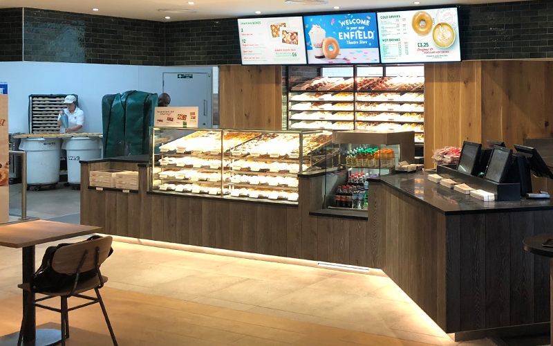 Interior of a donut shop with a display case of donuts, menu board, and a staff member.
