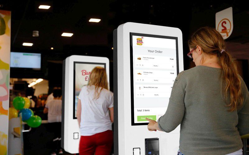 People ordering food at a kiosk in a restaurant. One person touches the screen, while another waits.