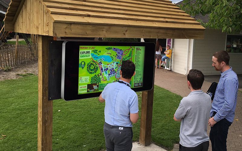 Three people looking at an interactive map on a digital kiosk outdoors. Wooden structure with a green map.