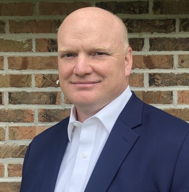 A man in a suit and white shirt is standing in front of a brick wall.