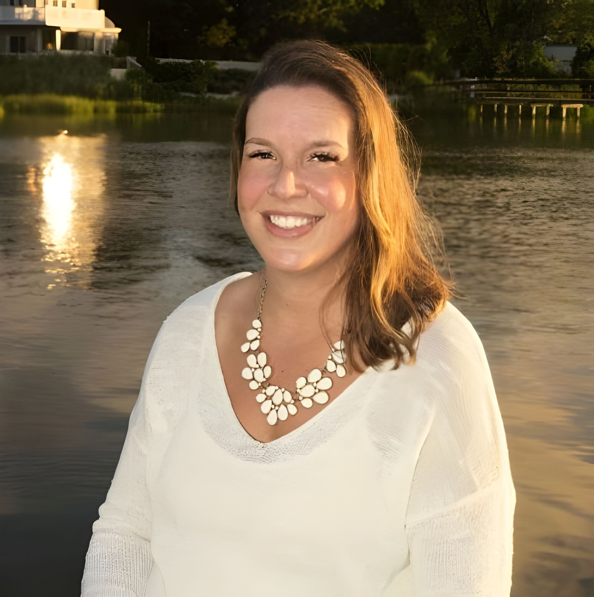A smiling person in a cream-colored sweater and a white statement necklace, posing outdoors by a calm lake at sunset.