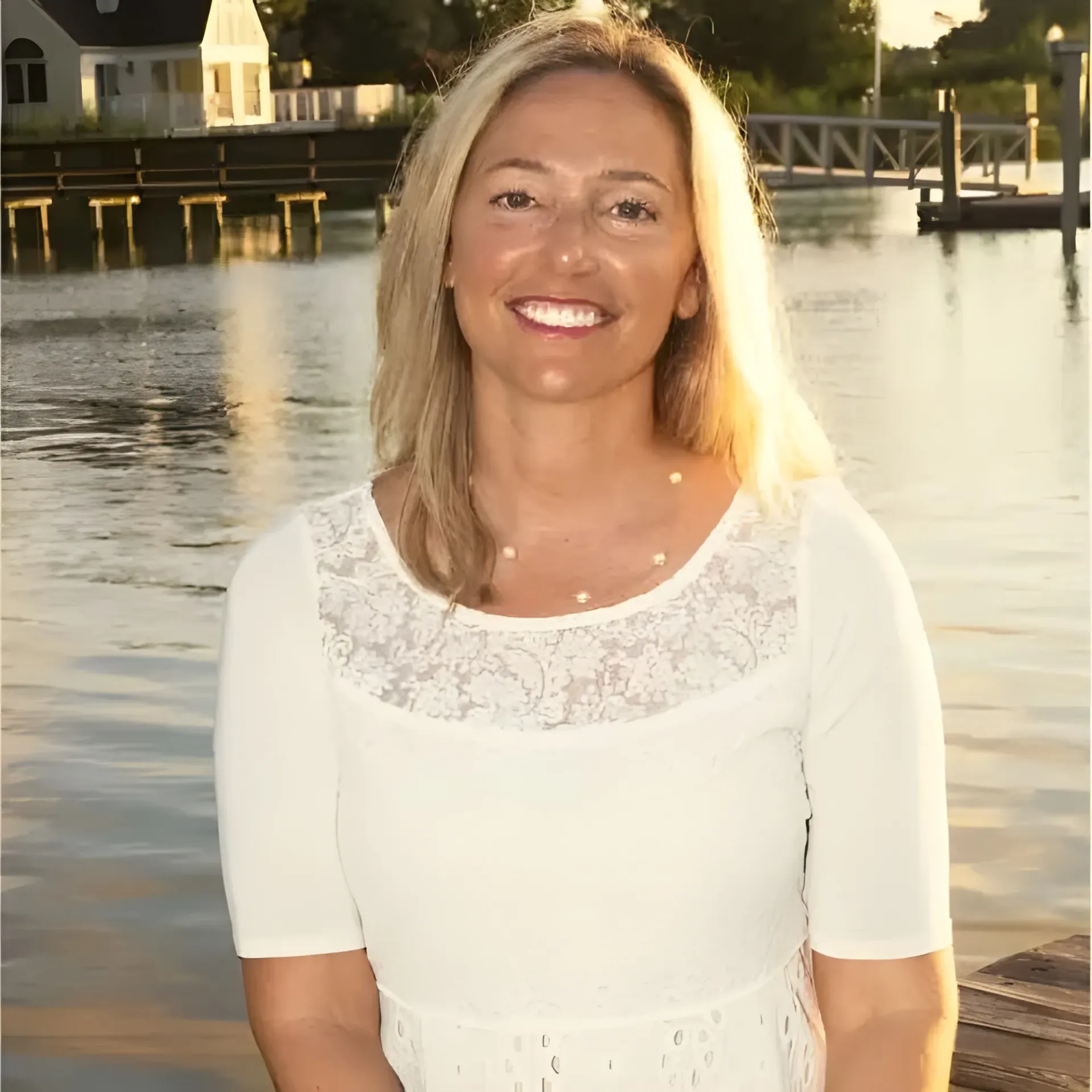 A smiling woman wears a white lace-top dress against a backdrop of a calm lake and a wooden dock at sunset.