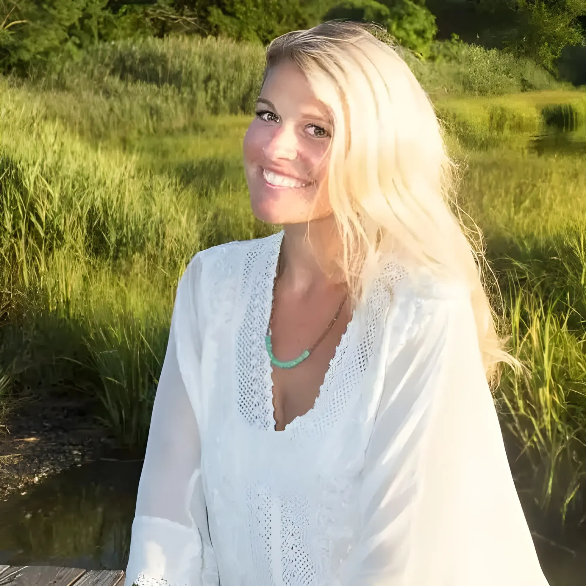 A smiling blonde person wearing a white lace-trimmed top and turquoise necklace, posed outdoors near green marsh grass.