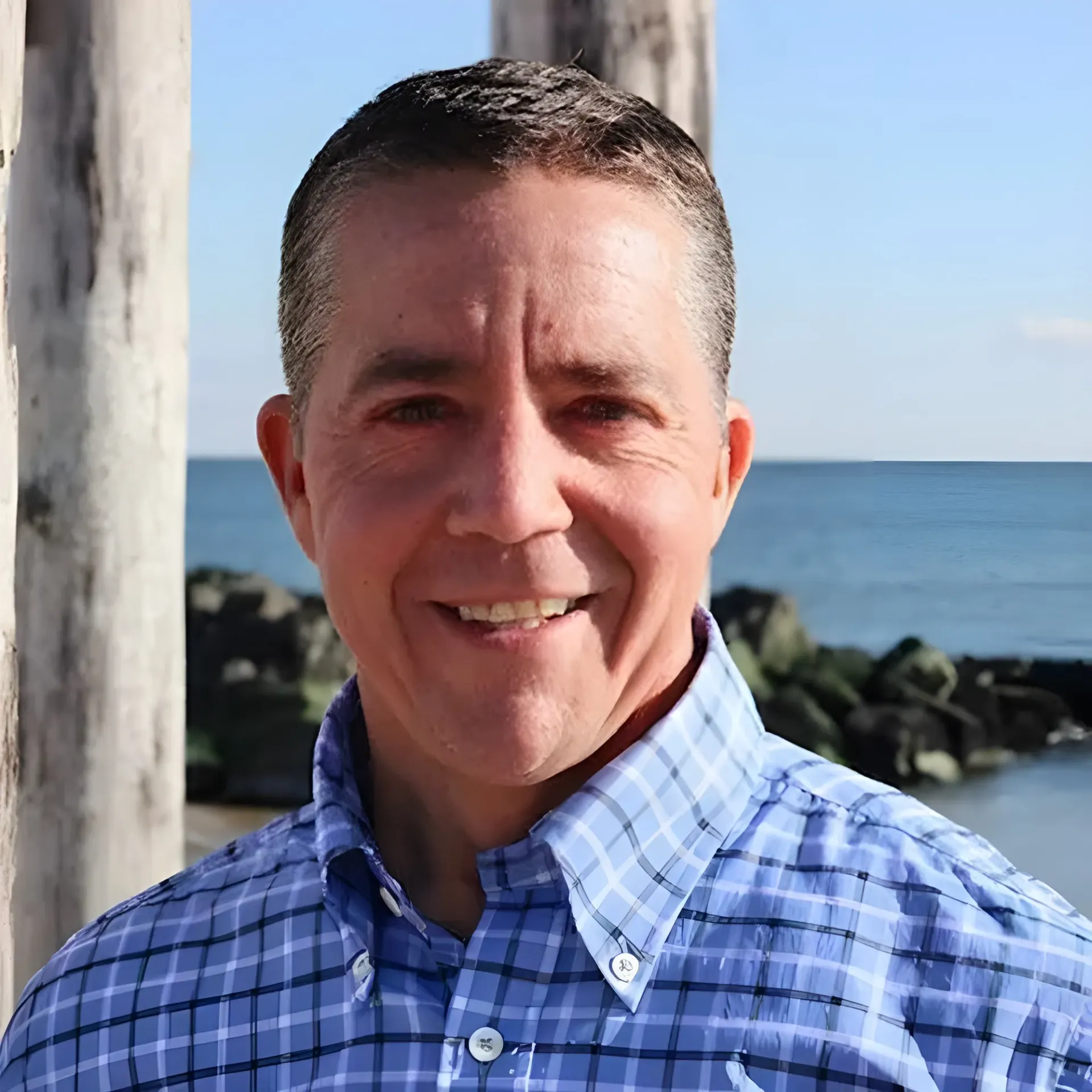A smiling person wearing a blue plaid button-down shirt, standing outdoors with a shoreline and ocean in the background.