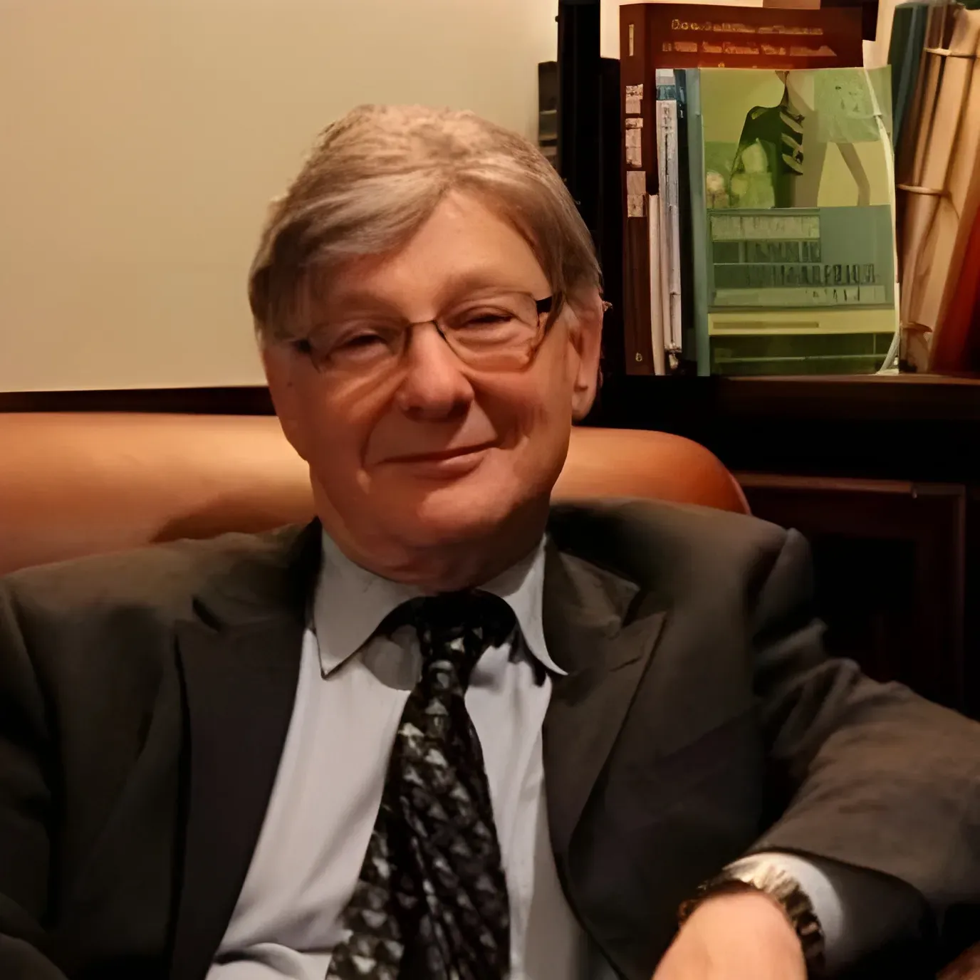 A person wearing a suit and tie smiling while seated in front of a bookshelf containing medical textbooks.