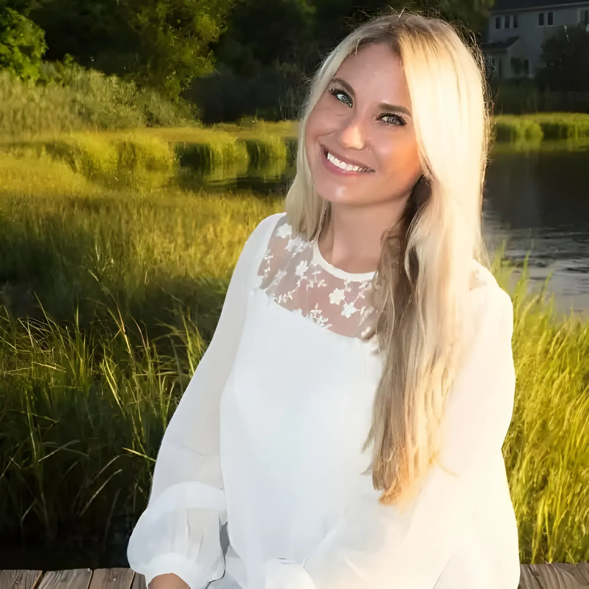 A blonde person with a warm smile wearing a white blouse, posed outdoors in front of tall grass and water at sunset.