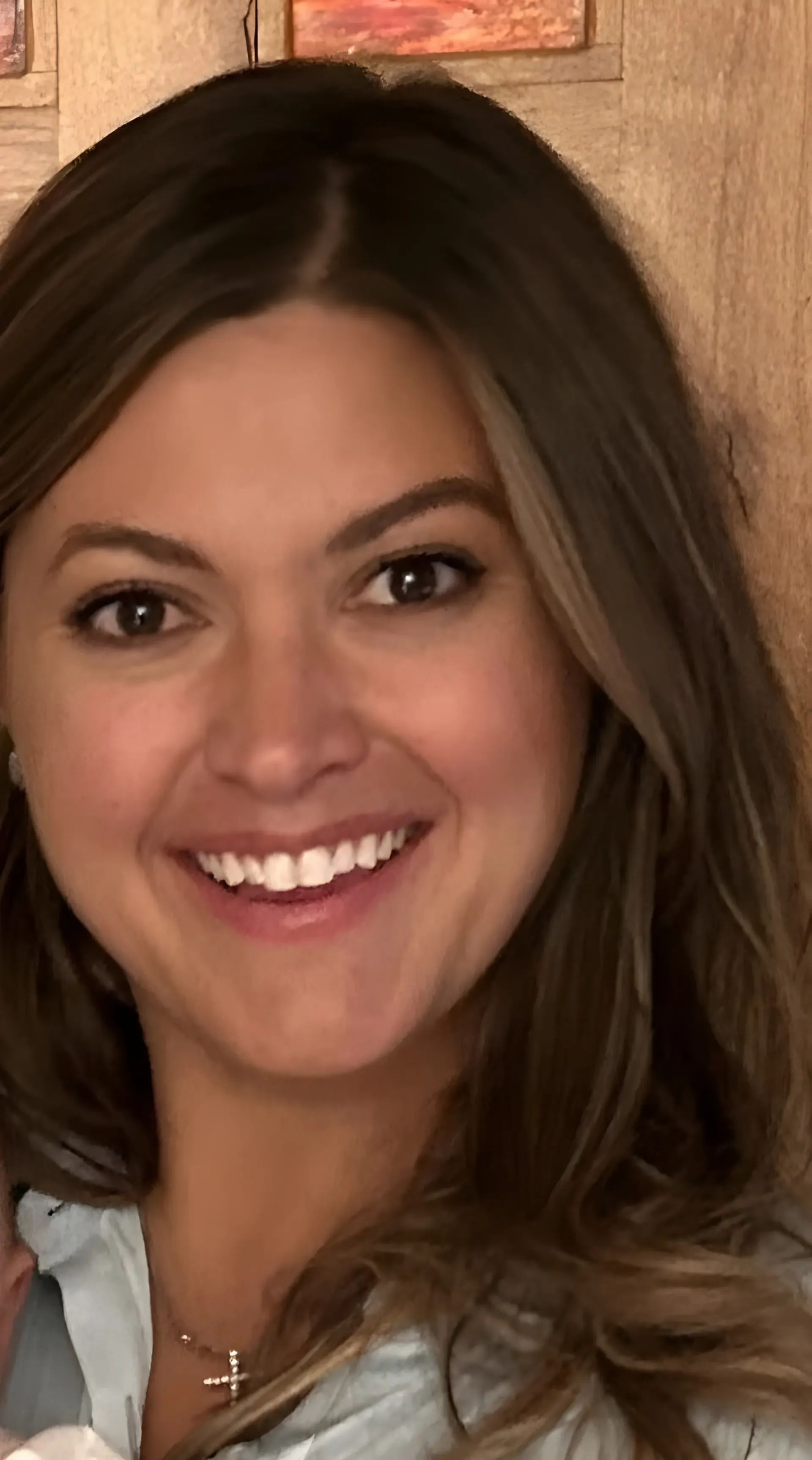 Close-up portrait of a smiling person with shoulder-length brown hair wearing a light blue top and a small cross necklace.
