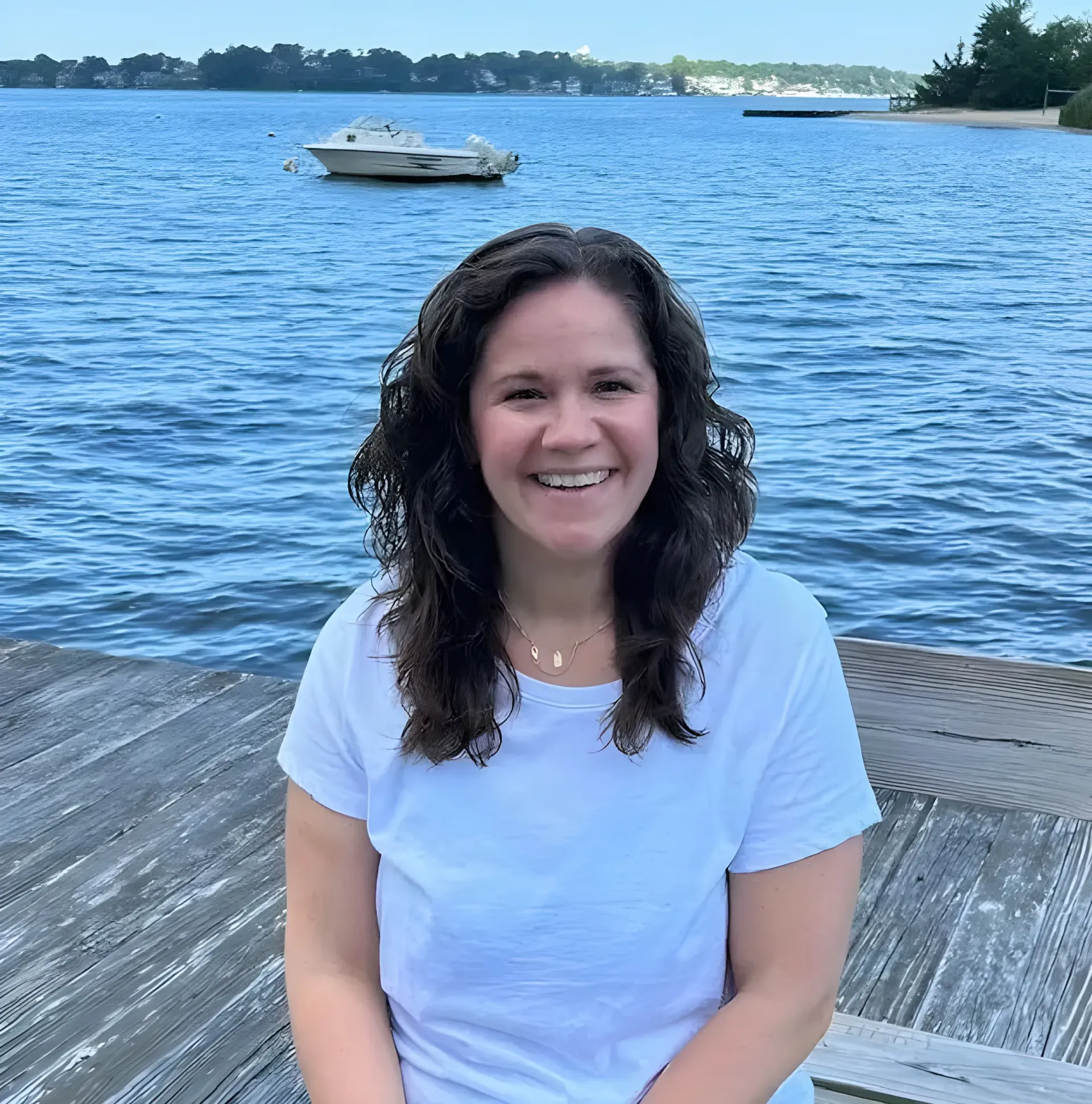 A smiling person with wavy brown hair wearing a white t-shirt, sitting on a wooden dock with a boat on the water behind.