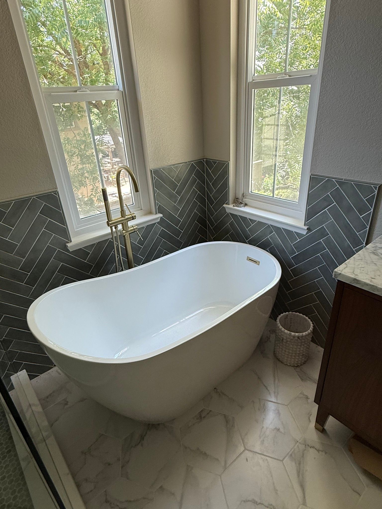 Bathroom with white bathtub, gold faucet, windows, and patterned tile.