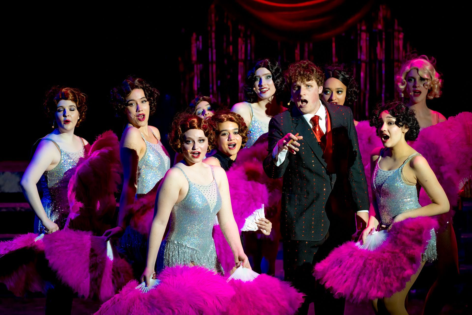 Professional headshot of a person with dark hair in a black top, posing with hands under chin against a dark reflective background.