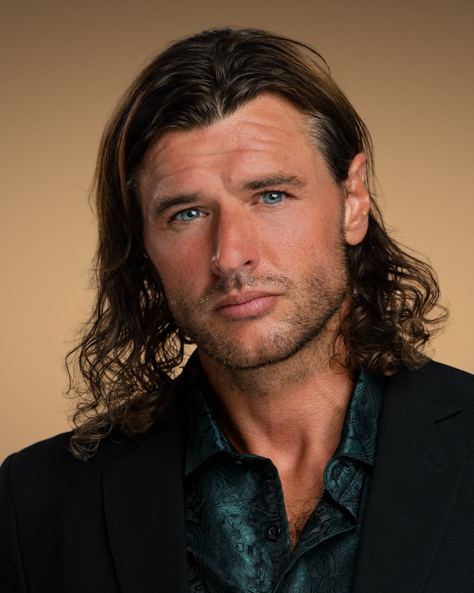 Professional Headshot of Lauren Mendez with shoulder-length, layered brown hair wearing a black blazer over a light gray top. A delicate gold necklace with a rectangular pendant is visible. The background is solid black, creating a professional studio portrait setting.