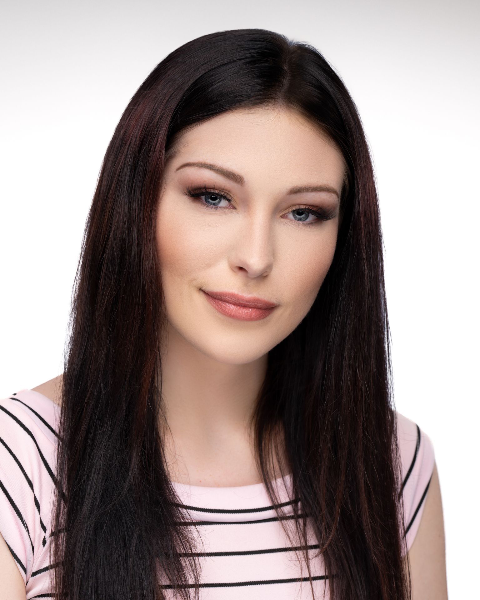 Portrait of a person with long straight dark hair wearing a light pink top with black stripes, photographed against a clean white studio background.